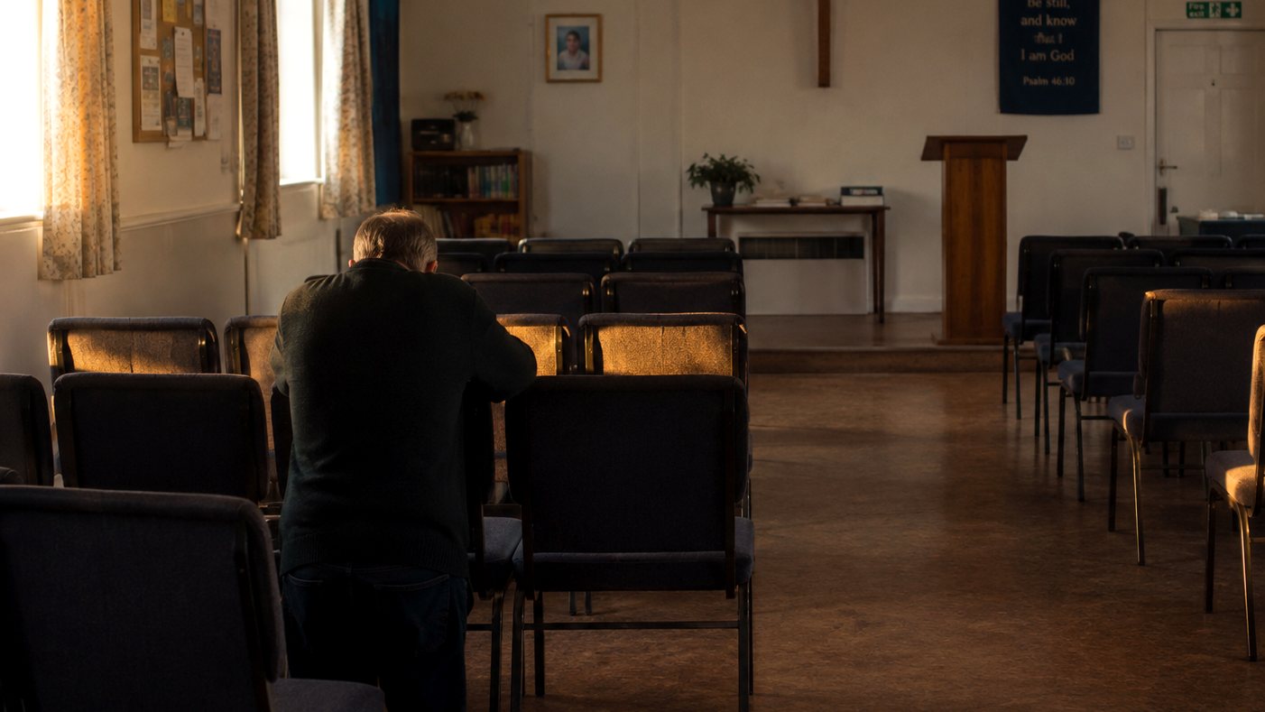 A man kneeling beside a chair in a quiet church hall early on a Sunday morning, rows of empty chairs around him in low golden light
