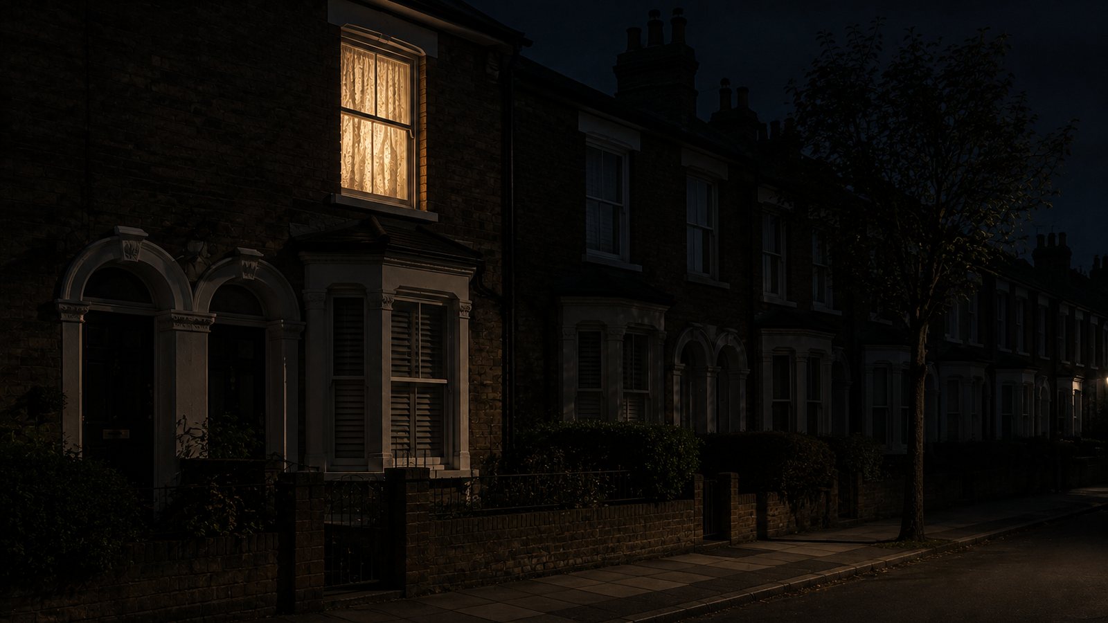 A single lit window in a terraced British street at night, warm lamplight behind the curtain while the rest of the street is dark